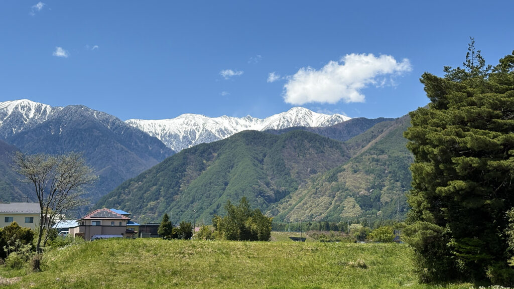 春の北アルプスの風景。青空の下、白く雪化粧した険しい山々と、緑豊かな山麓が広がり、その手前には新緑に包まれた田園地帯と民家が点在するのどかな風景。