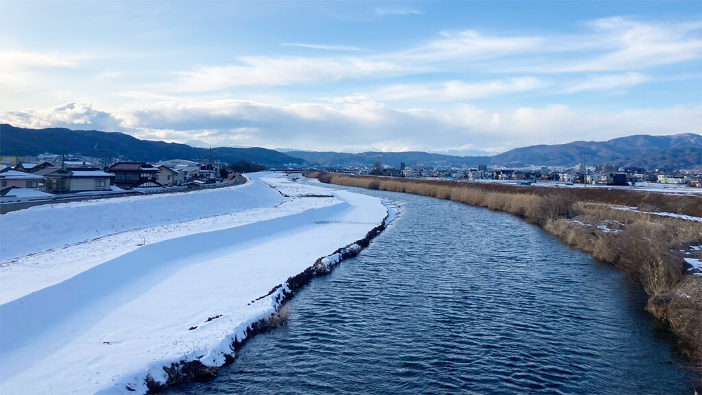 冬の長野の川辺風景。雪に覆われた堤防と青く澄んだ川が並び、遠くには連なる山々と雲が広がる広大な空が印象的な冬の光景。