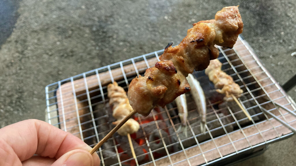 炭火で焼かれた香ばしい焼き鳥。手に持たれたジューシーな焼き鳥の串、その背景には七輪の上で焼かれる他の串や魚が見える。庭での食事風景。